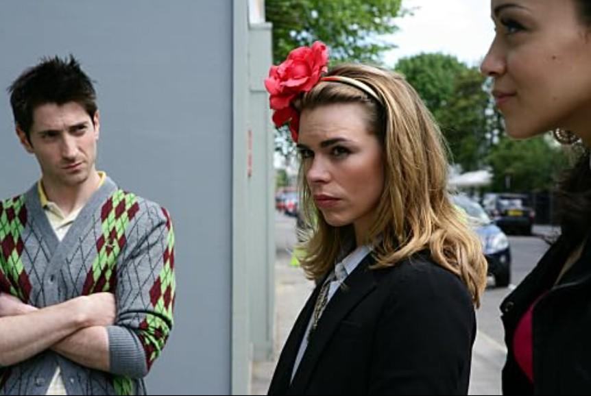 Drei Personen stehen draußen. Ein Mann links, eine Frau mit Blume im Haar in der Mitte.