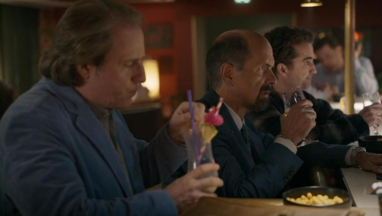 Drei Männer sitzen an einer Bar, einer hält ein buntes Getränk, ein anderer trinkt Champagner.