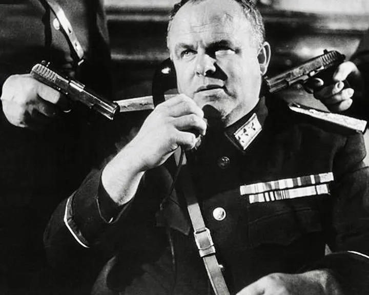 Mann in Uniform hält ein Telefon, zwei Pistolen sind auf ihn gerichtet.