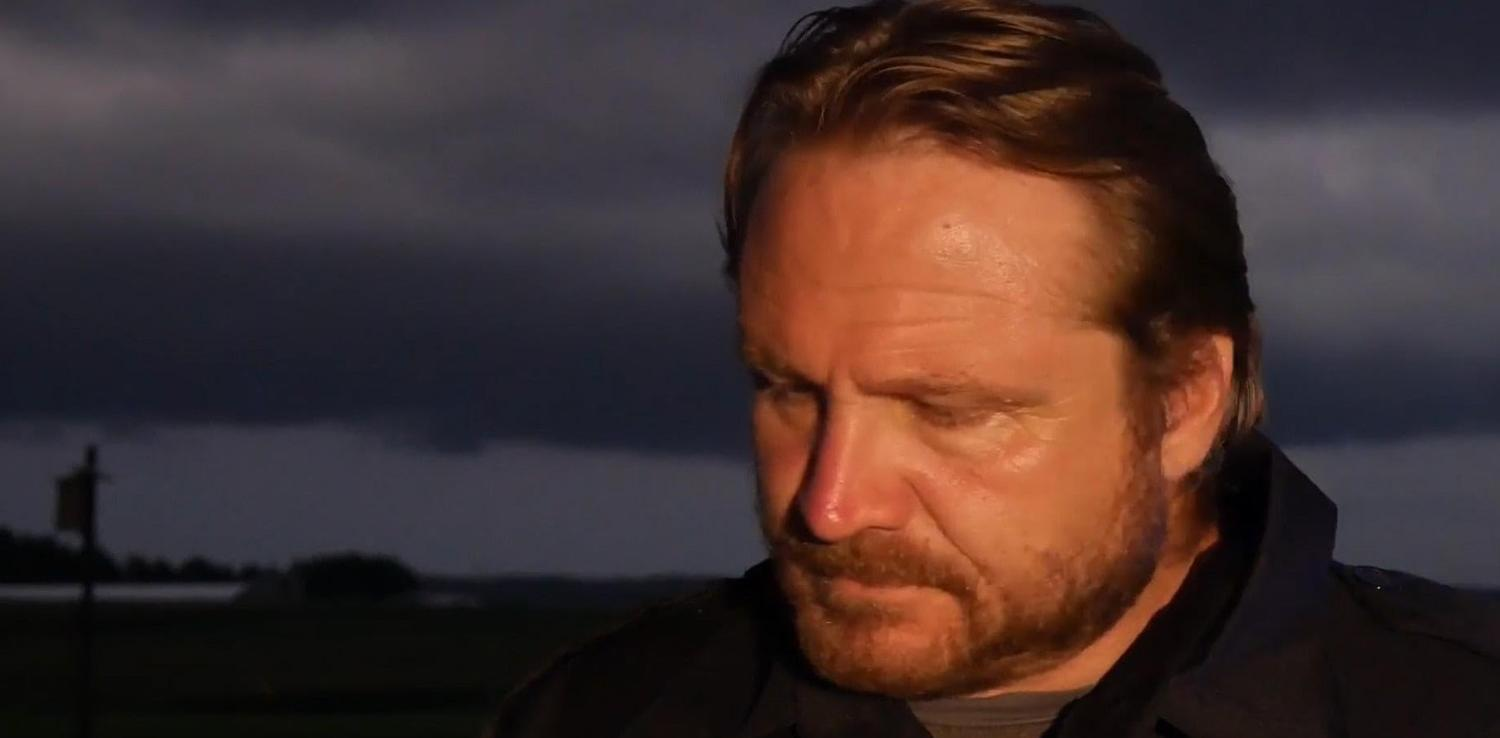 Ein Mann mit Bart blickt nach unten vor einem dunklen, dramatischen Himmel.