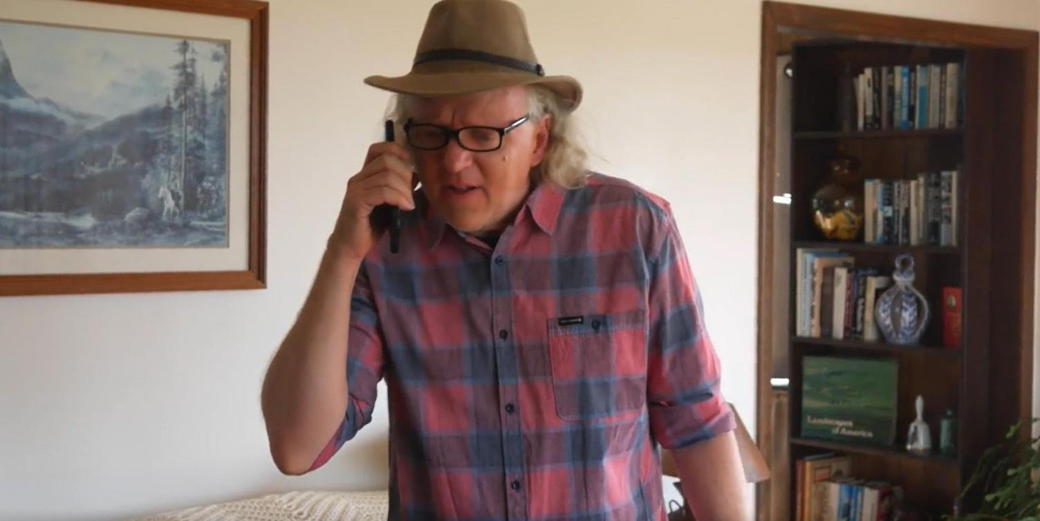 Mann mit Hut und Brille telefoniert, kariertes Hemd, gerahmte Kunst und Bücherregal.
