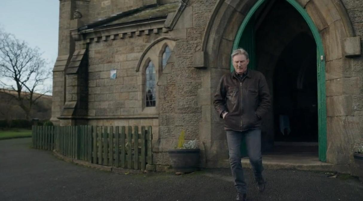 Ein Mann in Lederjacke verlässt eine Steinkirche durch einen grünen Torbogen.