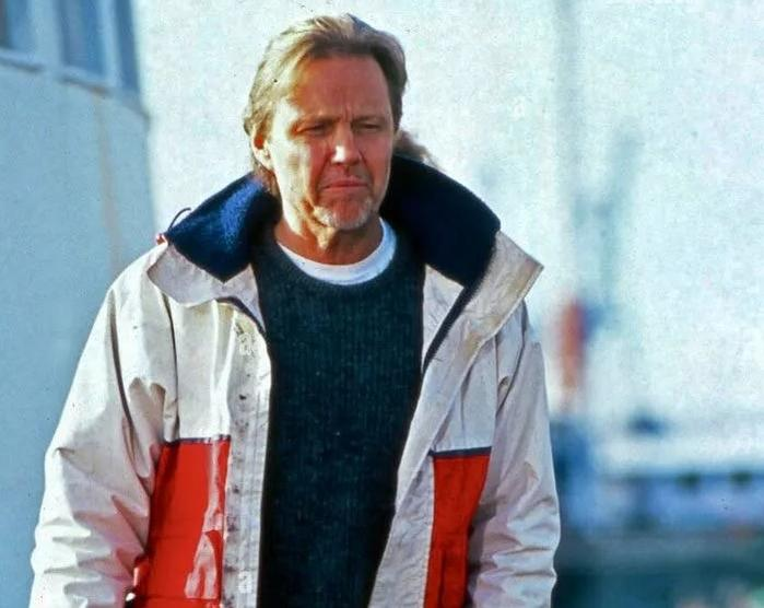 Ein Mann mit blonden Haaren trägt einen dunkelgrünen Pullover und eine mehrfarbige Jacke.