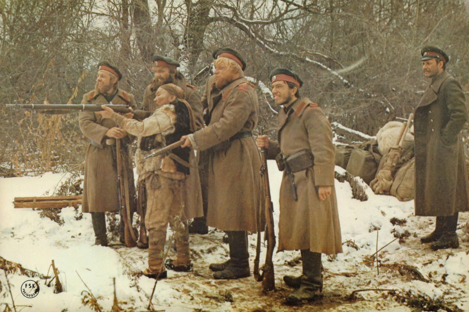 Fünf Soldaten in Wintermänteln mit Gewehren im Schnee, eine Person in der Mitte reicht ein Gewehr.