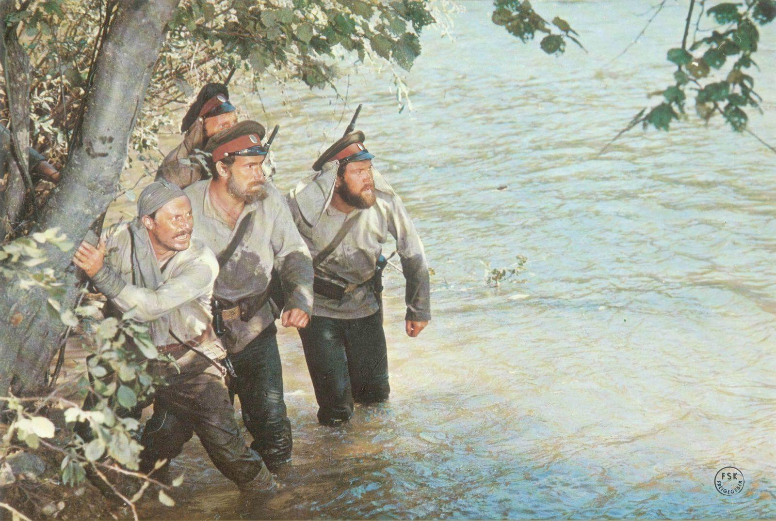 Vier Soldaten in Uniform, die durch flaches Wasser waten, im Schatten eines Baumes.