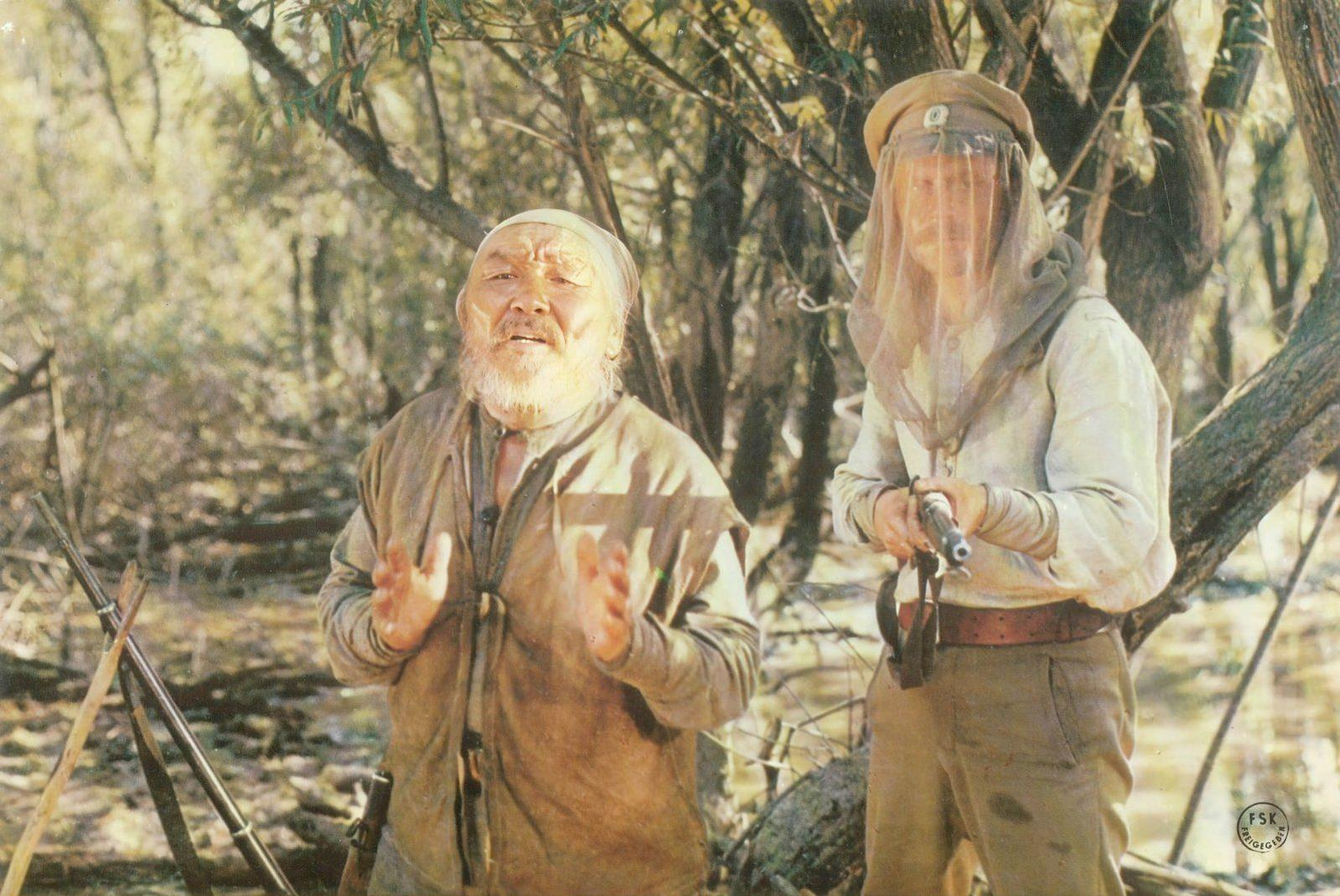 Ein alter Mann mit Bart steht neben einem Mann mit Schleier und Gewehr im Wald.