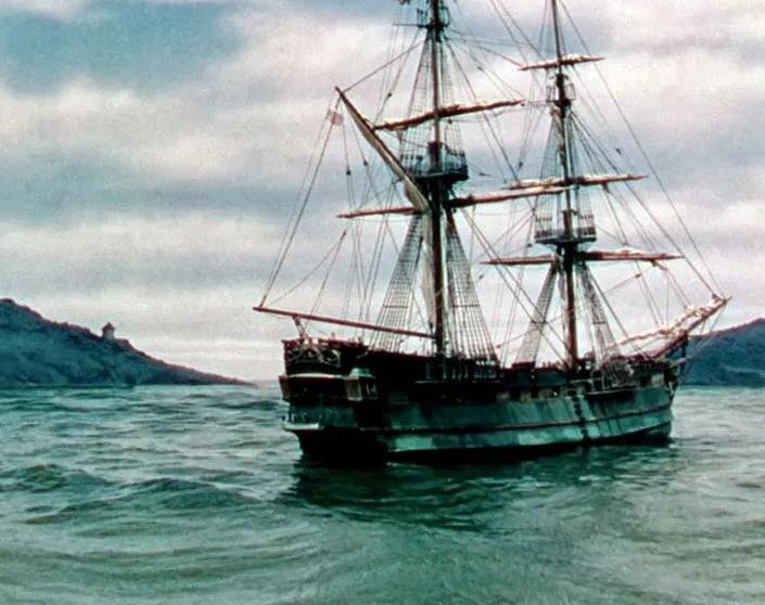 Ein Segelschiff mit mehreren Masten fährt auf dem Meer, mit einer bewaldeten Küste im Hintergrund.