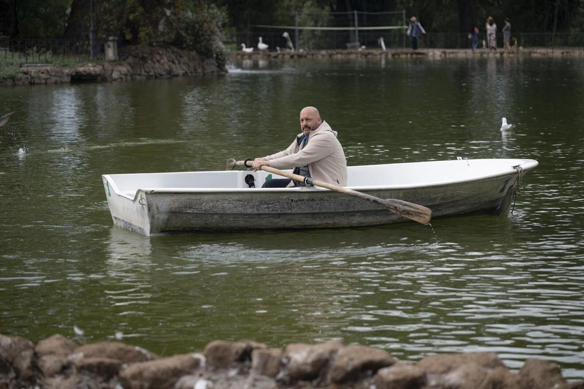 Ein Mann rudert in einem weißen Boot auf einem Teich. Im Hintergrund sind Enten und Menschen zu sehen.