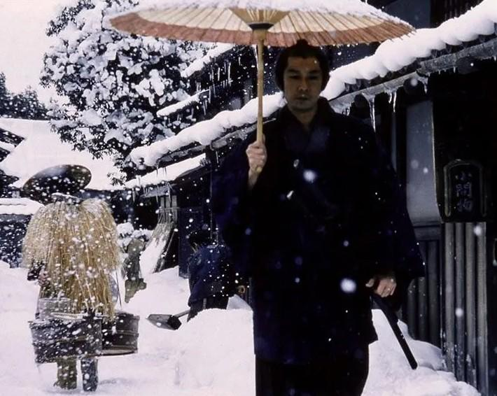 Ein Mann mit Regenschirm geht durch tiefen Schnee vor traditionellen Häusern.