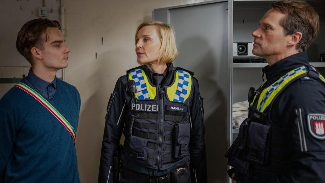 Drei Personen, zwei Polizisten in Uniform und ein Mann in einem blauen Pullover, stehen zusammen.