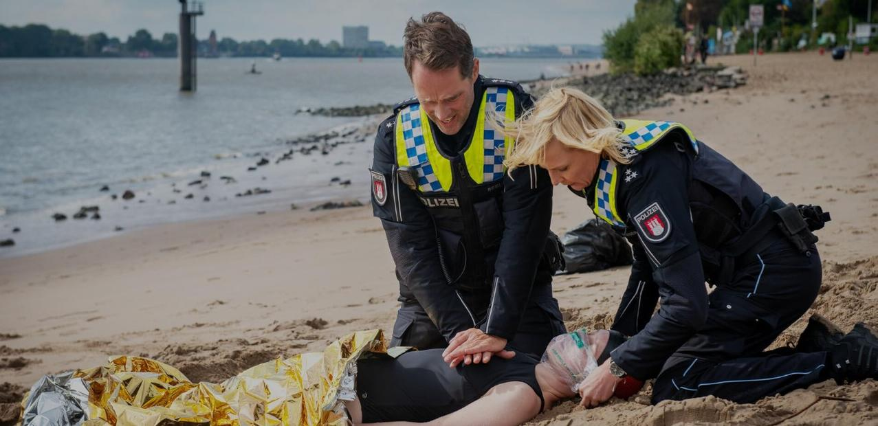 Polizisten auf einem Strand, die Erste Hilfe leisten. Eine Person liegt auf dem Sand, bedeckt mit Folie.