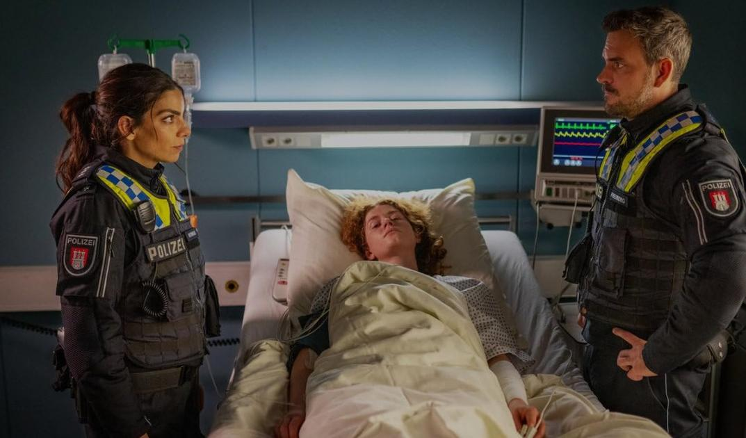 Zwei Polizisten stehen neben einem Patienten im Krankenhausbett.