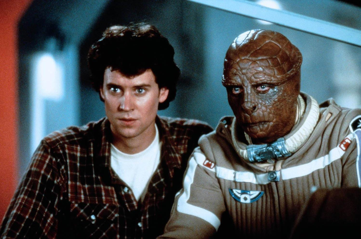 Ein Mann mit dunklem lockigem Haar trägt ein kariertes Hemd neben einem Alien mit brauner Schuppenhaut.