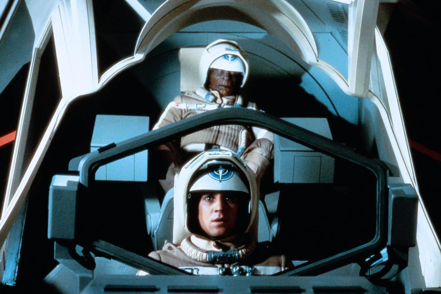 Zwei Piloten in Helmen und beigen Anzügen sitzen in einem futuristischen Cockpit.