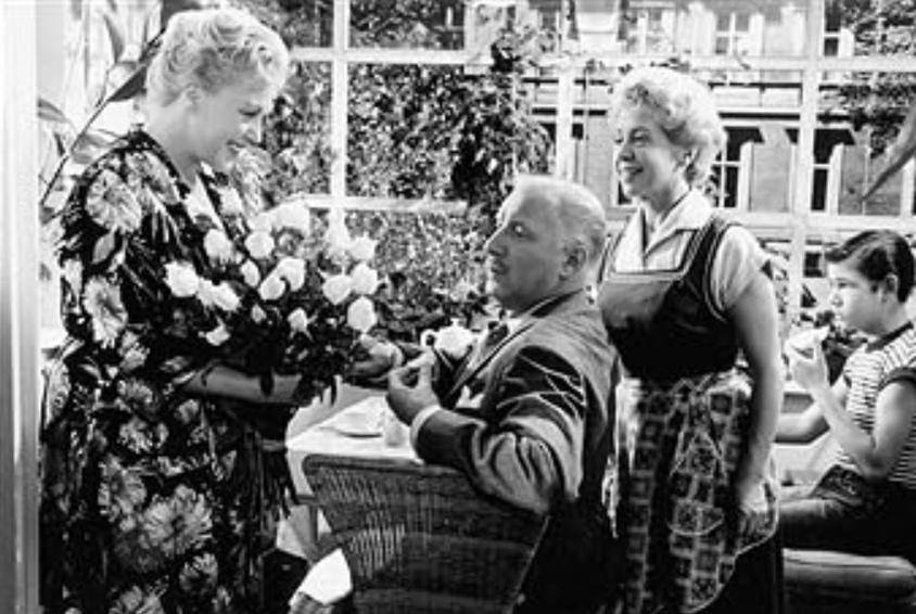 Vier Personen sitzen oder stehen im Freien; eine Frau gibt Blumen.