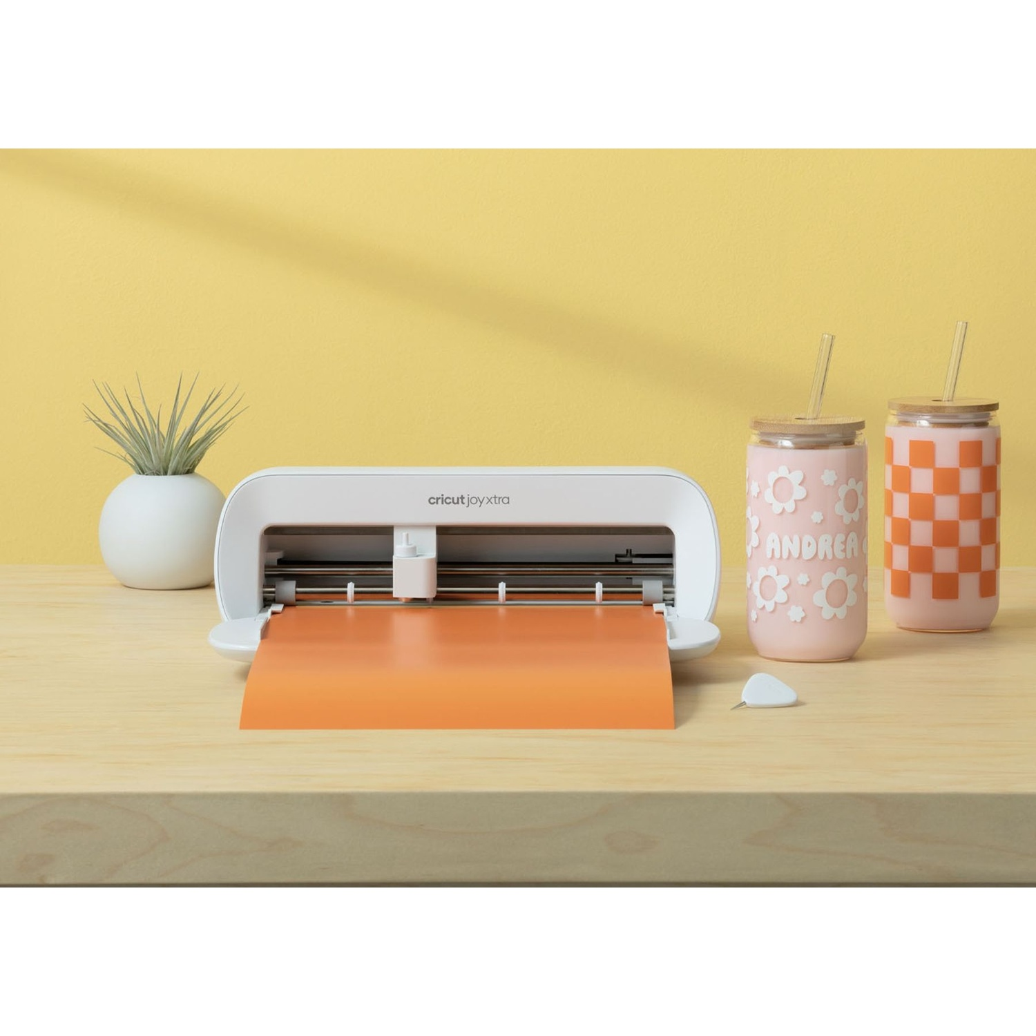 Una macchina da taglio Cricut Joy Xtra bianca con vinile arancione, due tazze e una pianta.