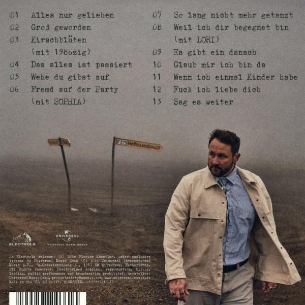 Mann mit beige Jacke steht vor zwei Wegweisern. Trackliste im Hintergrund.