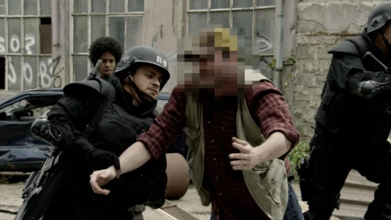 Polizisten in schwarzer Rüstung nehmen einen Mann in kariertem Hemd fest.