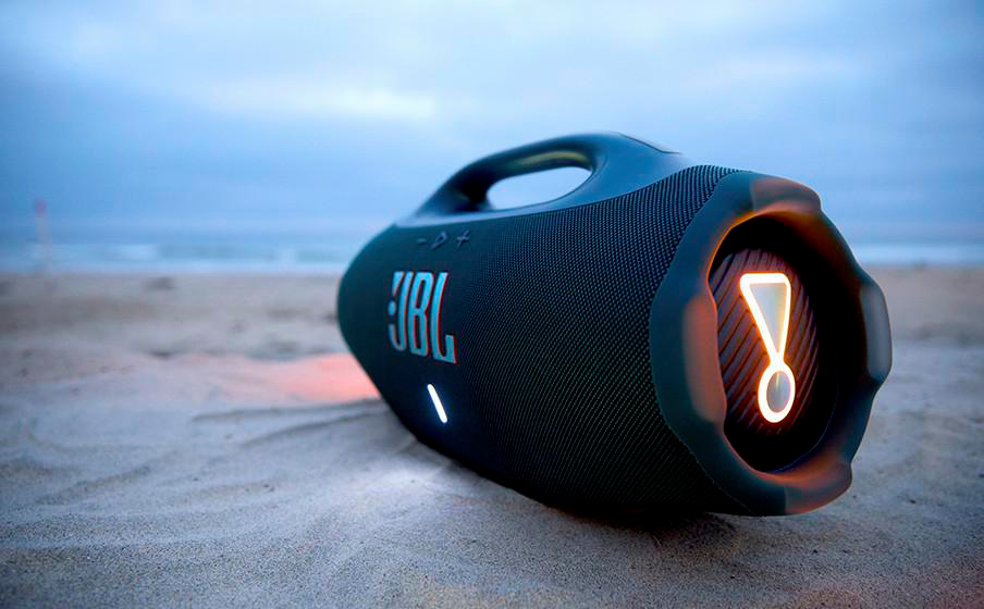 Ein schwarzer JBL-Lautsprecher liegt am Strand, mit dem Meer im Hintergrund.