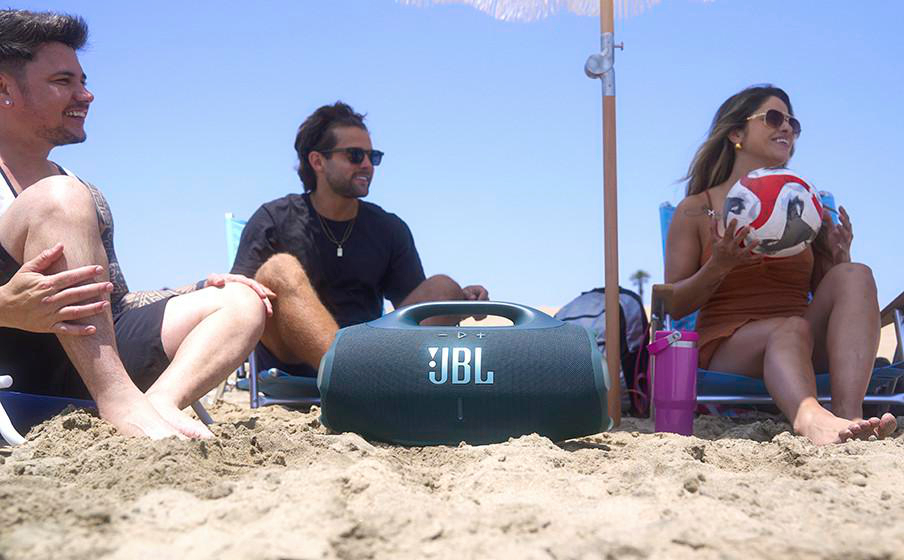 Drei Personen sitzen am Strand auf Liegestühlen. Ein JBL-Lautsprecher steht im Sand.