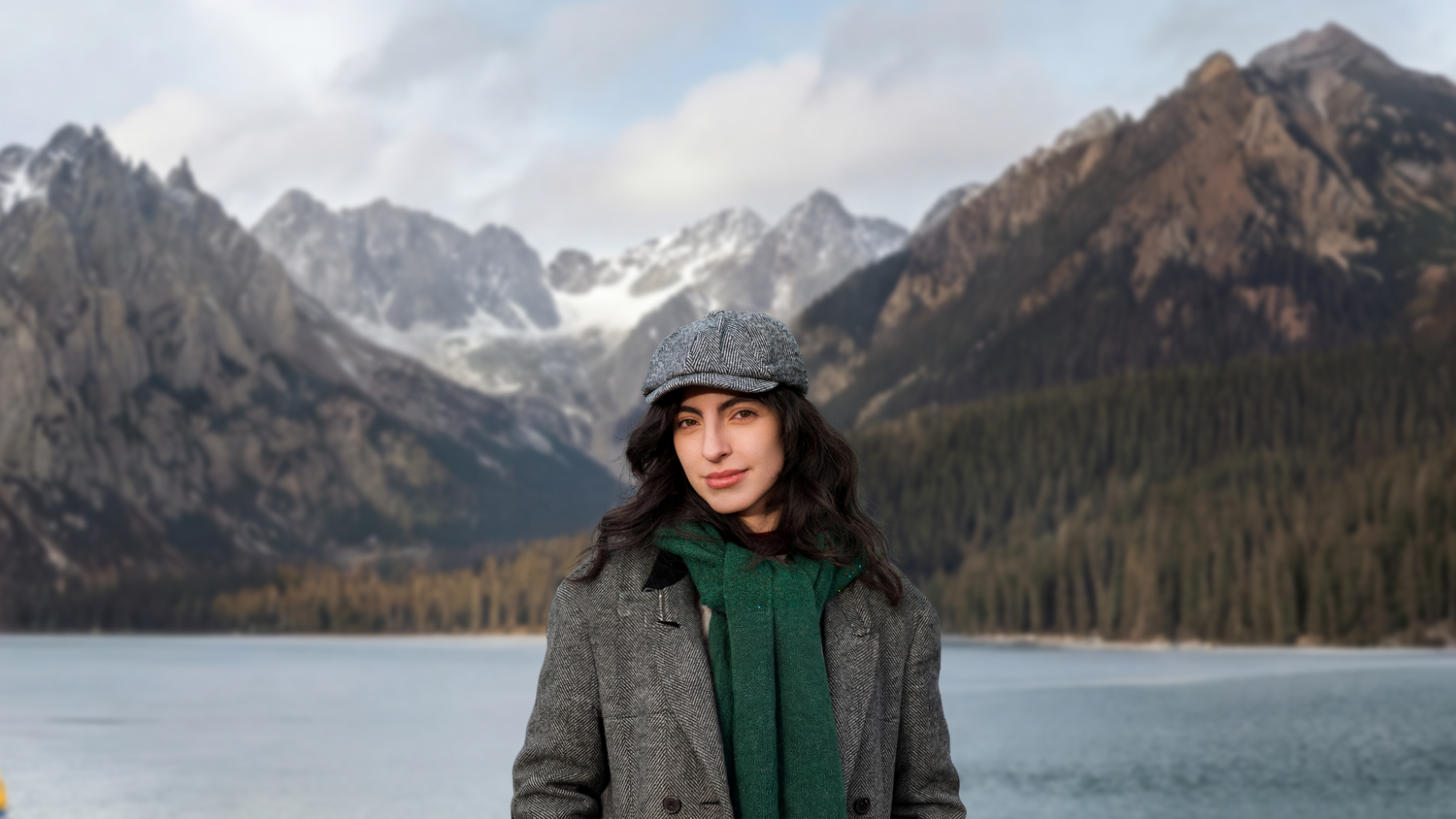 Una mujer con gorra y bufanda se alza ante un lago y montañas nevadas.