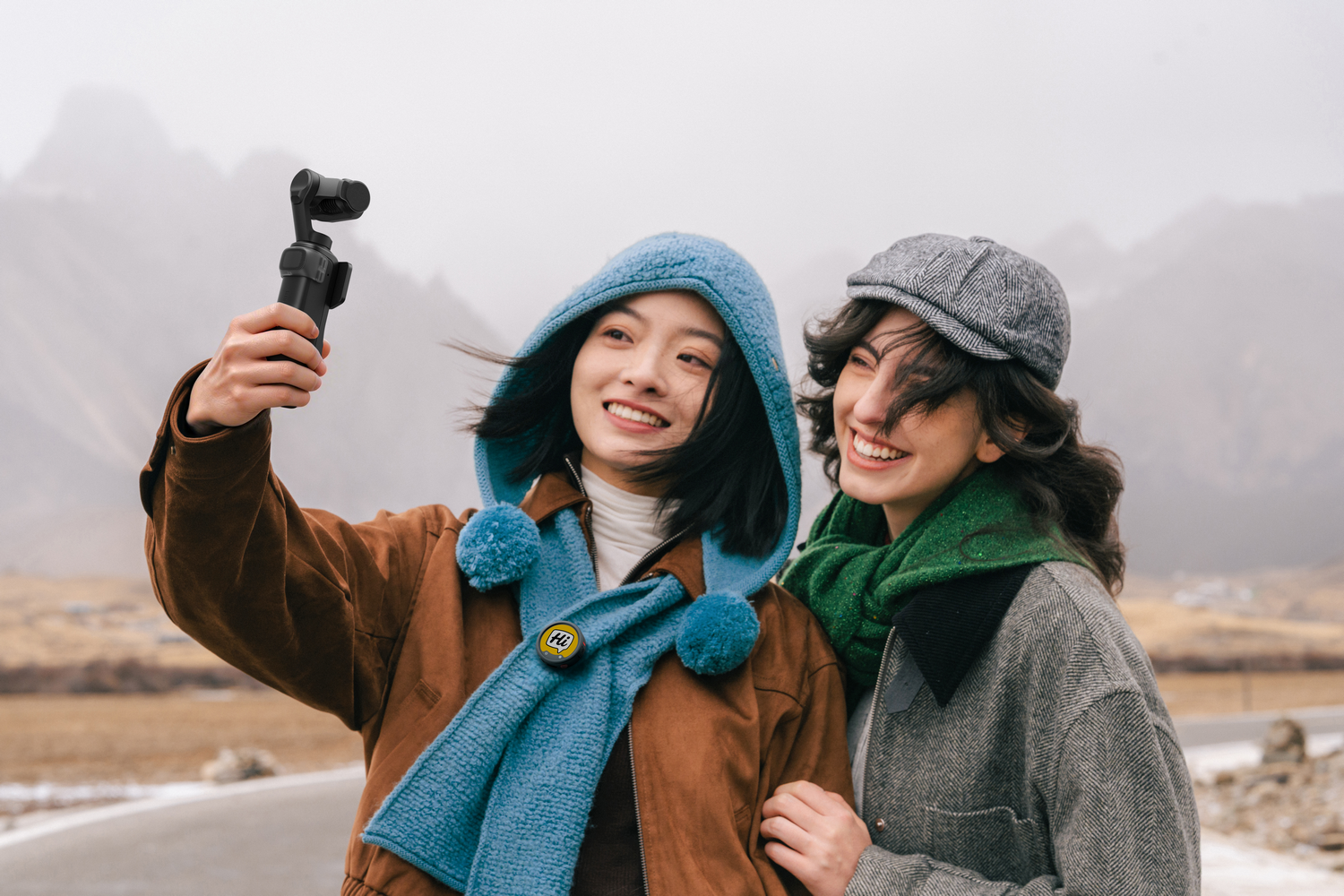 Dos mujeres sonríen para una selfie con un gimbal en un paisaje montañoso y neblinoso.