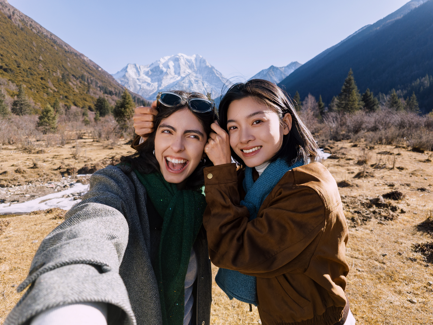 Dos mujeres sonrientes con ropa abrigada se toman una selfie en un paisaje montañoso con parches de nieve.