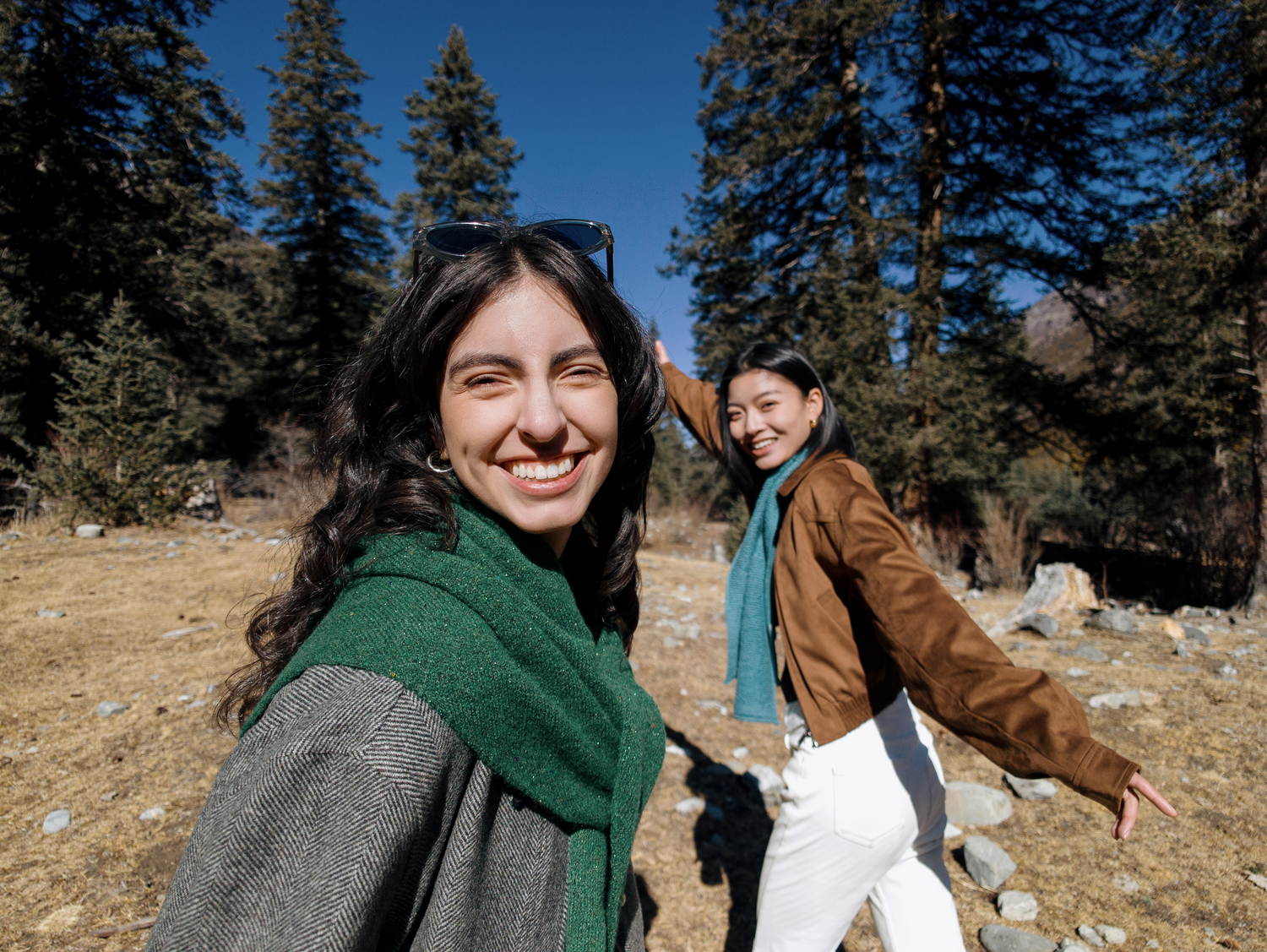 Dos mujeres sonriendo en un bosque. La mujer del frente lleva una bufanda verde y un abrigo gris.