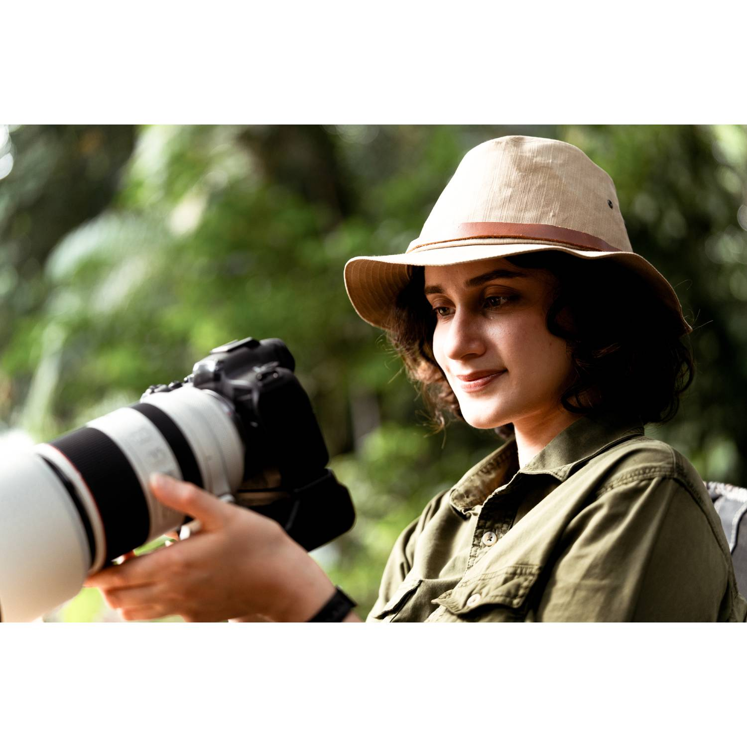 Una mujer con sombrero safari sostiene una cámara grande con lente zoom, al aire libre.