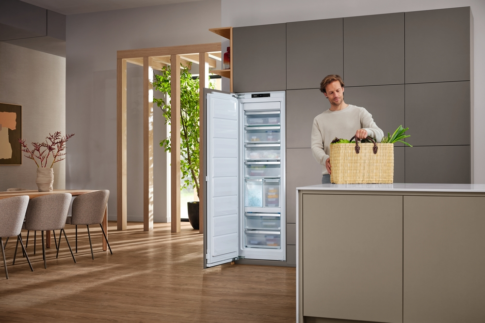 Mann stellt Einkaufskorb mit Gemüse in eine offene, moderne Küche neben einen Kühlschrank.