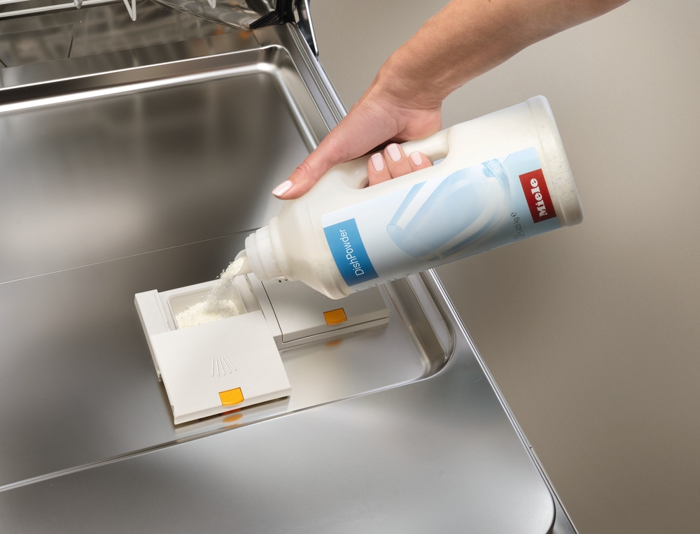 Eine Hand gießt Spülpulver aus einer weißen Flasche in einen Spülmittelspender.