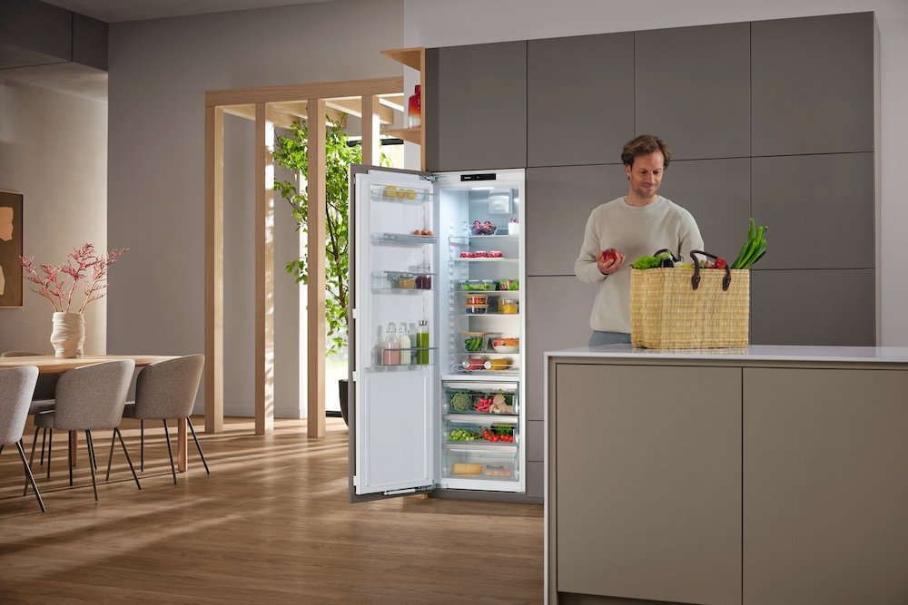 Mann mit Apfel steht neben geöffnetem Kühlschrank und Einkaufskorb mit Gemüse.