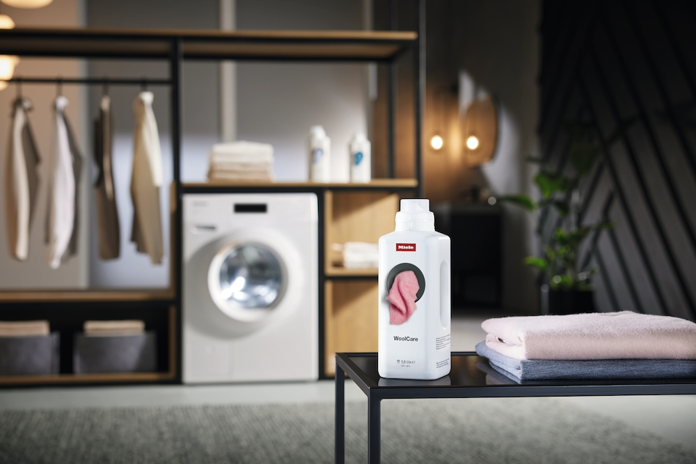 Miele WoolCare Waschmittelflasche steht neben gestapelten Pullovern in einer Waschküche.