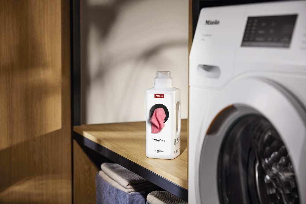 Eine Miele WoolCare Waschmittelflasche steht neben einer Waschmaschine.