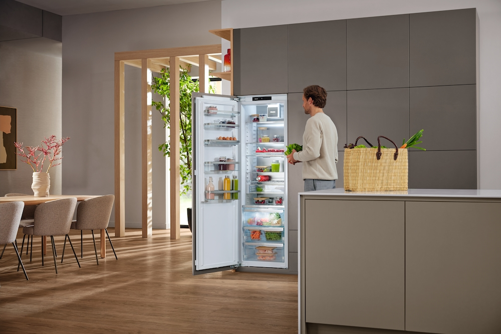 Mann legt Salat in offenen Kühlschrank. Auf der Theke steht eine Tasche mit Gemüse.