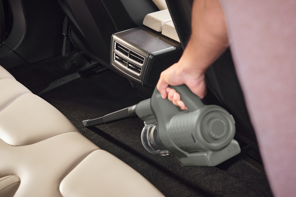 Hand saugt Auto-Boden mit grauem Staubsauger, beigefarbene Ledersitze.