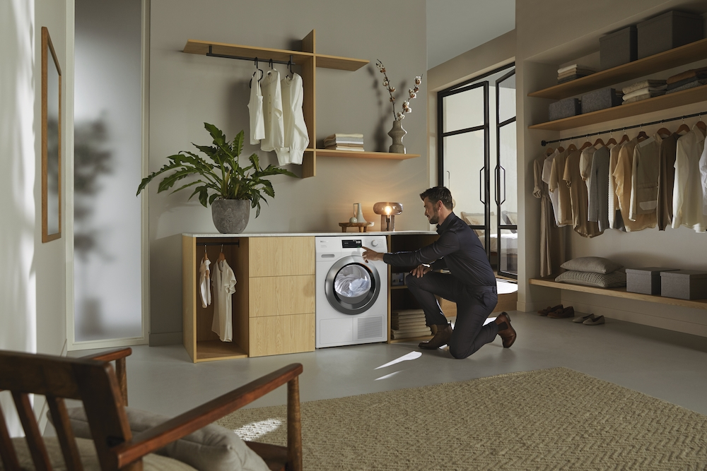 Mann bedient Waschmaschine neben Kleiderschrank mit Kleidung und Pflanze.