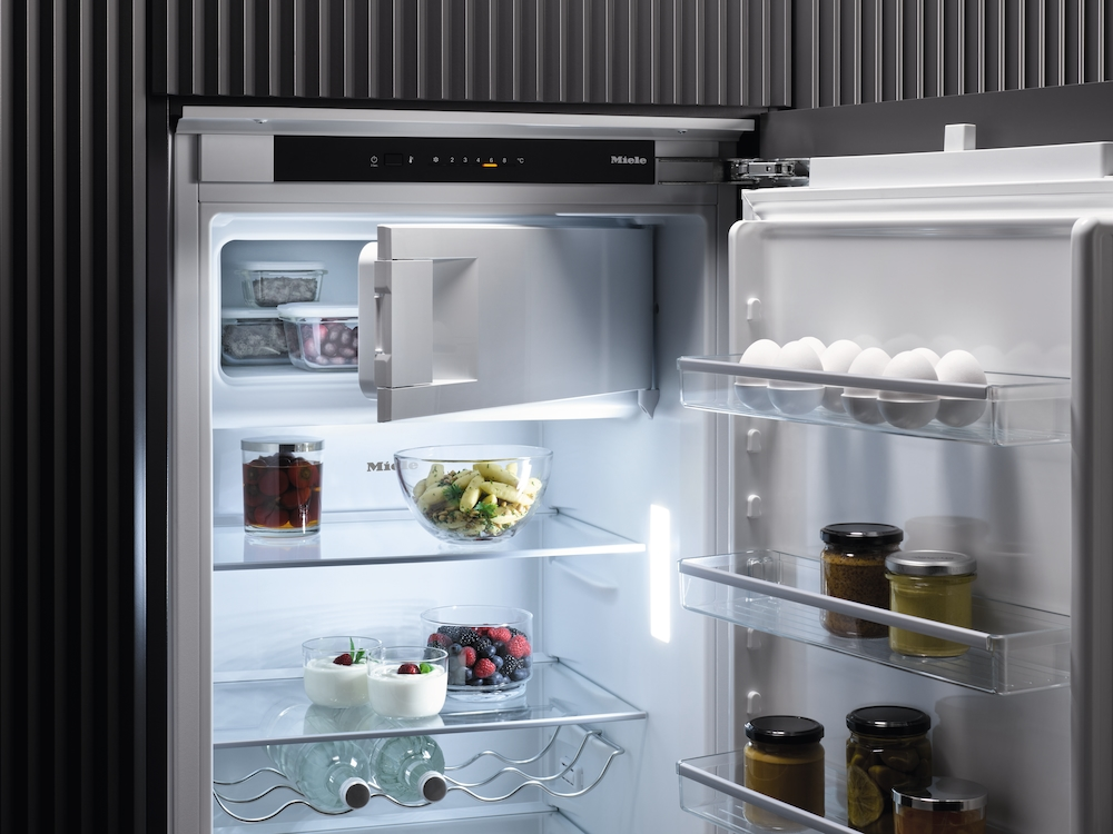 Offener Kühlschrank mit Lebensmitteln: Eier, Gläser, Obst- und Pastaschalen.