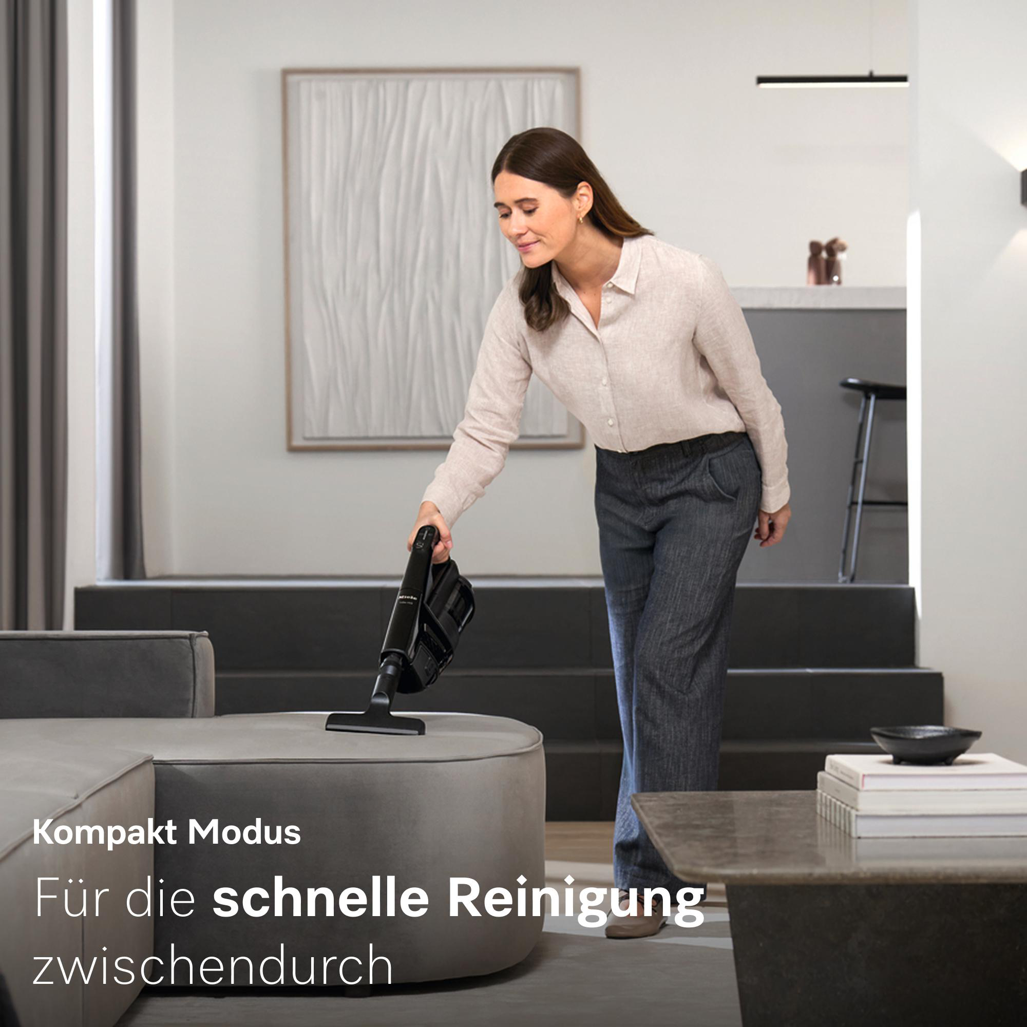 Frau benutzt schwarzen Handstaubsauger auf grauem Hocker im modernen Wohnzimmer.