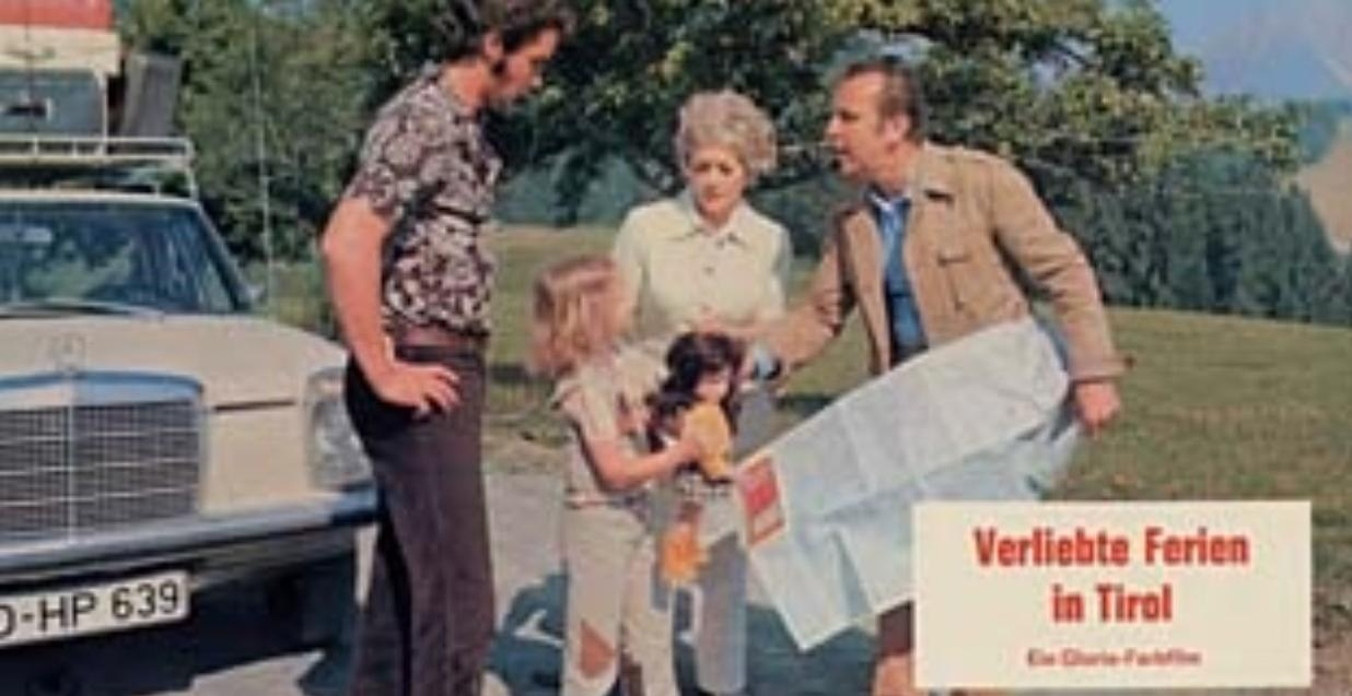 Familie mit Gepäck vor einem Auto, Karte wird gehalten. Berge im Hintergrund.