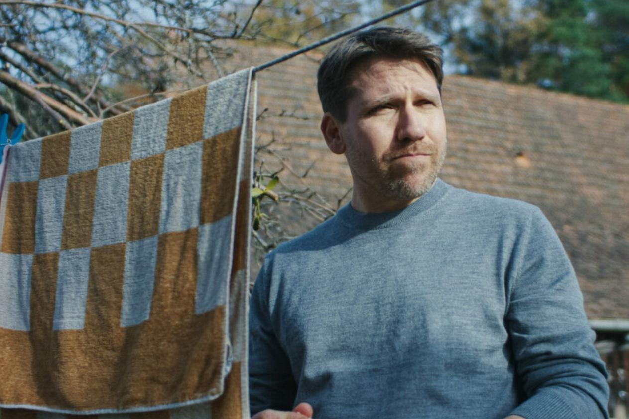 Ein Mann in einem grauen Pullover blickt nach rechts. Eine karierte Decke hängt daneben.