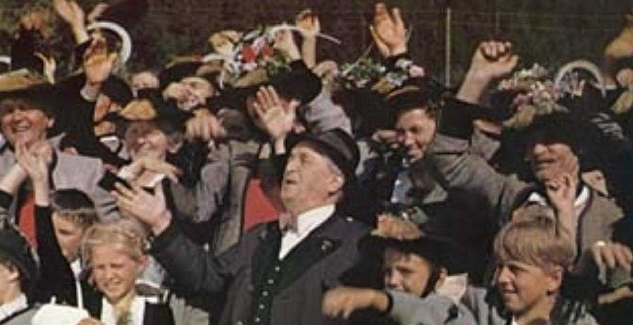 Eine Menge jubelnder Menschen mit Hüten und Blumen, einige winken.