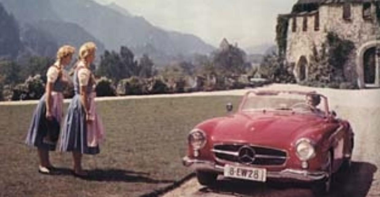 Zwei Frauen in Tracht stehen neben einem roten Oldtimer vor einem Schloss.