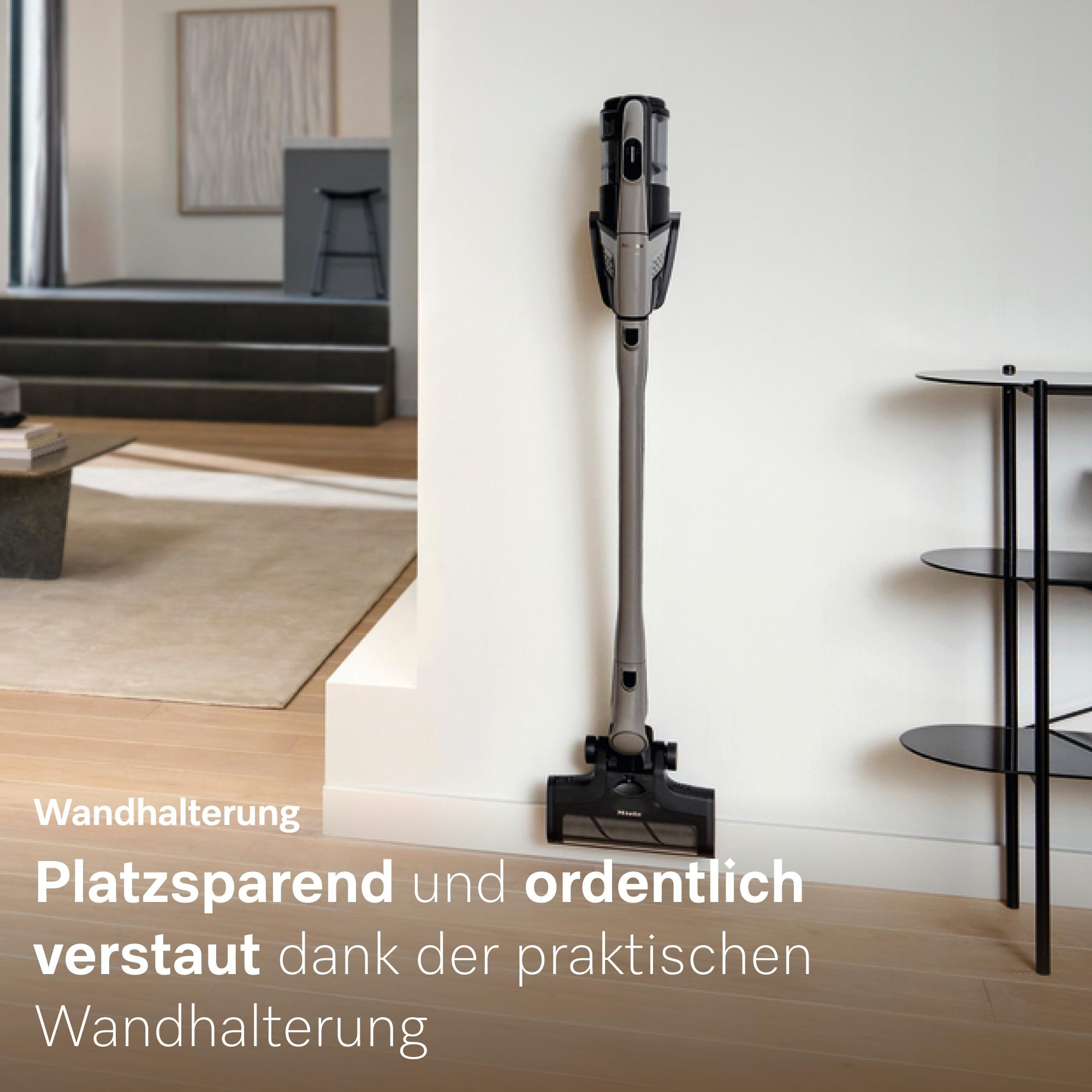 Ein grauer Akku-Staubsauger ist an einer weißen Wand montiert. Rechts steht ein schwarzes Regal.