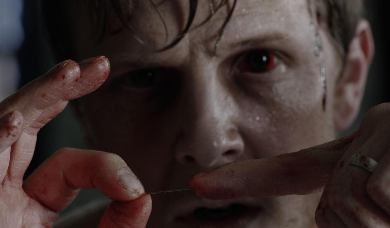 Ein Mann mit blutunterlaufenen Augen hält eine Nadel, seine Finger sind blutig.
