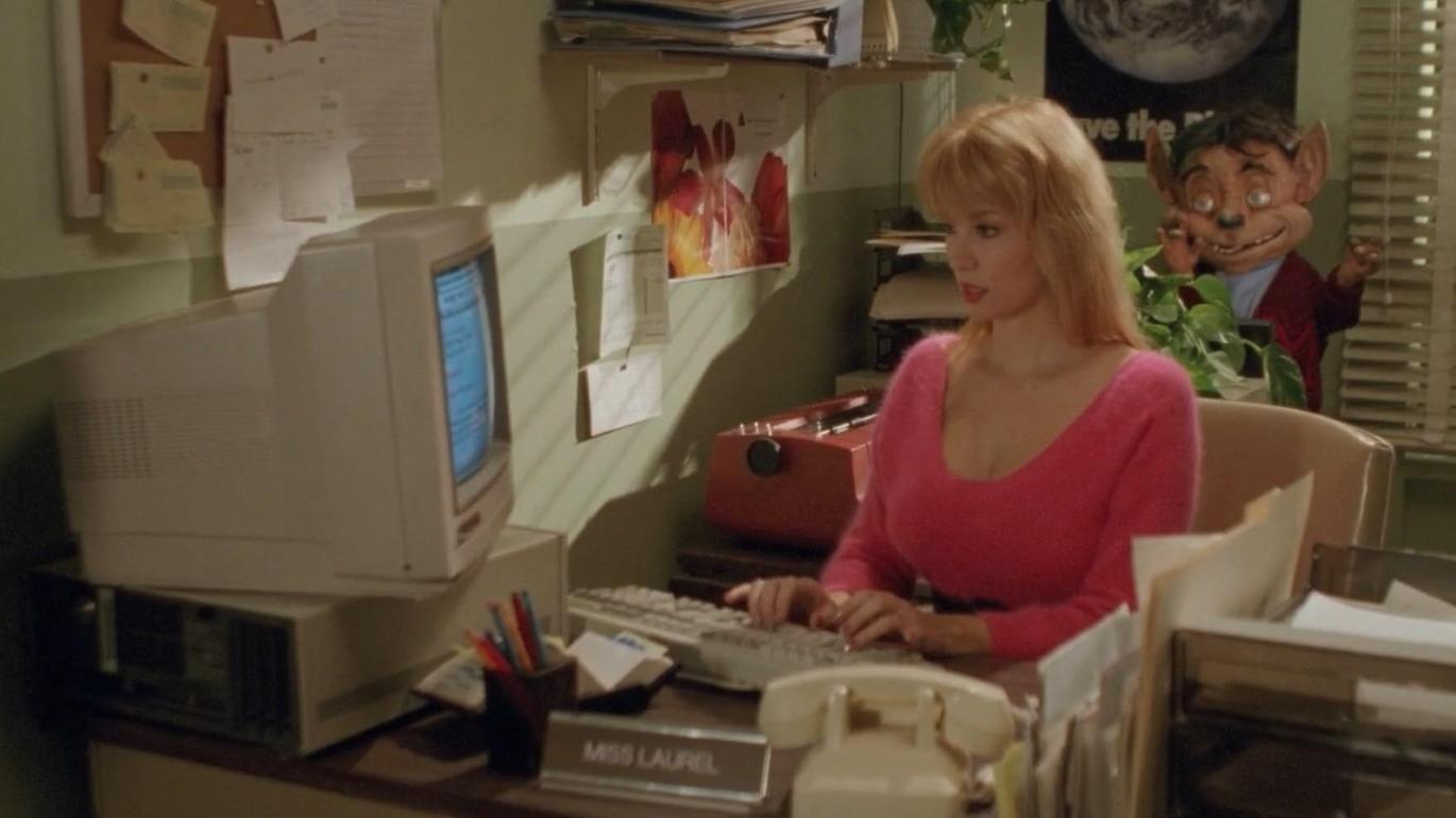 Eine Frau in einem rosa Pullover sitzt an einem Schreibtisch mit einem Computer. Ein Puppengesicht lugt hinter ihr hervor.