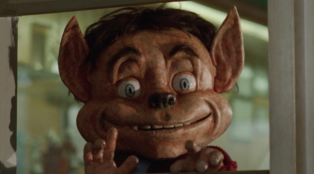 Ein grinsendes, groteskes Wesen mit großen Ohren und blauen Augen schaut hervor.