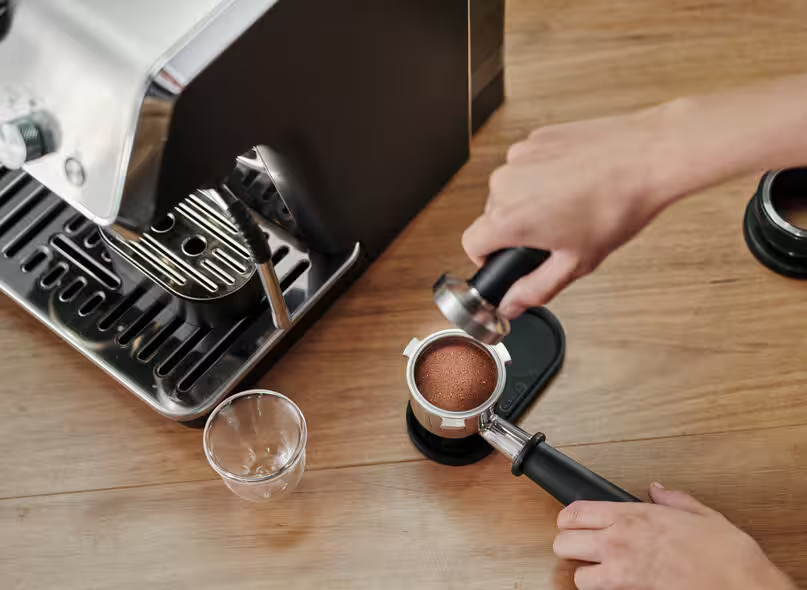 Ręka ubija kawę mieloną w portafiltrze obok ekspresu do kawy.