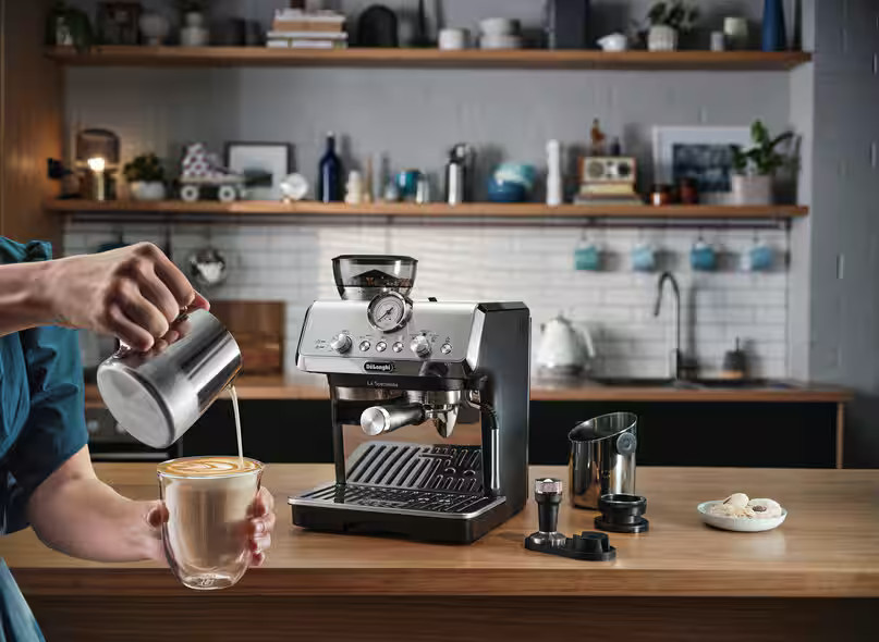 Osoba nalewa mleko do szklanki, tworząc latte art. Obok znajduje się ekspres do kawy.