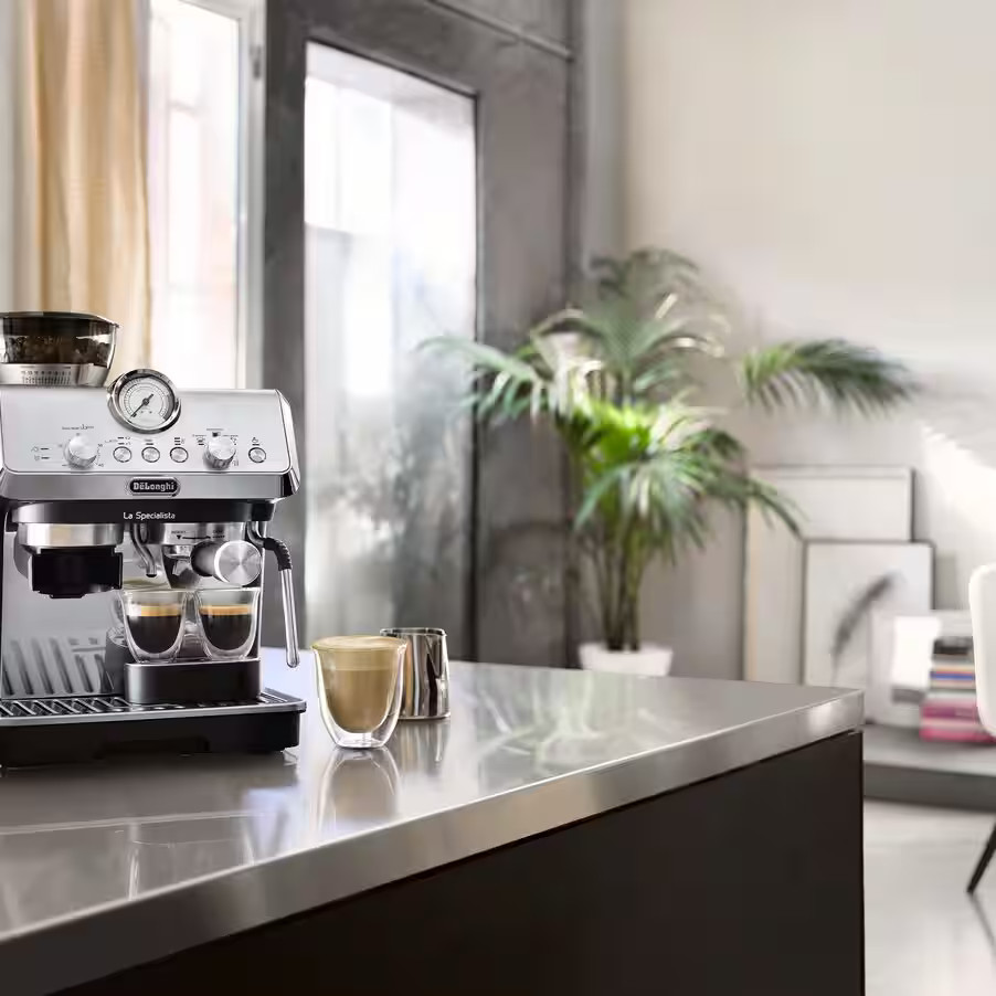 Srebrna maszyna do espresso z dwoma filiżankami kawy i rośliną w tle.